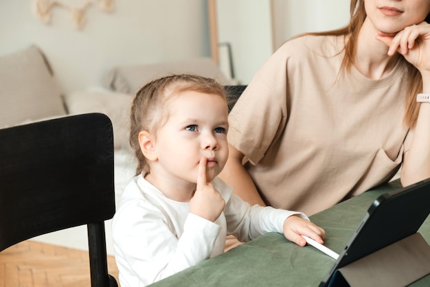 Ein kleines Mädchen denkt darüber nach, wie man zu Hause Aufgaben auf einem Tablet erledigt oder ein Spiel abschließt. Mama hilft bei den Hausaufgaben für den Kindergarten ihrer Tochter