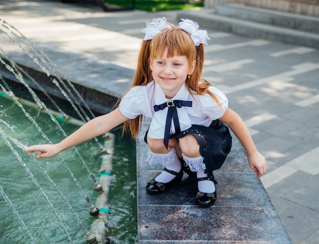 Ein kleines mädchen, ein grundschüler, spielt fröhlich in der nähe des brunnens. das kind erfrischt sich an einem heißen tag. ruhe nach dem schulunterricht.