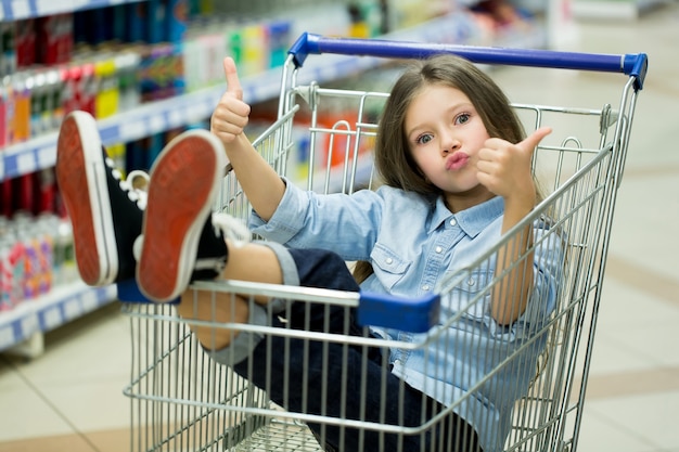 Ein kleines Mädchen fährt mit einem Karren in einem Supermarkt und zeigt mit dem Finger nach oben.