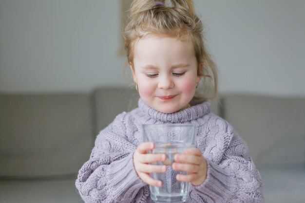 Ein kleines Mädchen hält ein Glas Wasser und trinkt. Gemütlich.