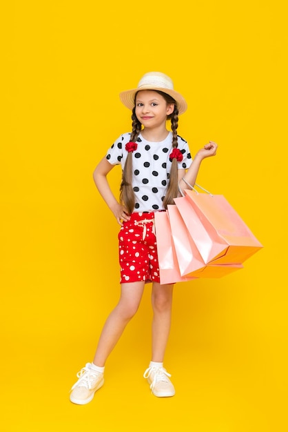 Ein kleines Mädchen hält Einkaufstüten Ein Kind in einem Sommerhut und Shorts ist mit Kindereinkäufen beschäftigt Ein glückliches Kind in vollem Wachstum auf einem gelb isolierten Hintergrund