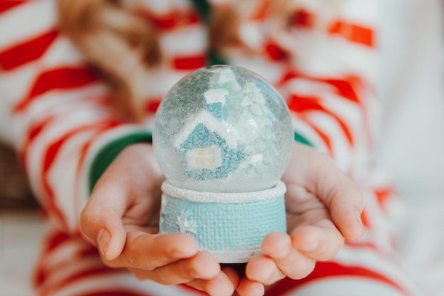 Ein kleines Mädchen im Pyjama und einer Weihnachtsmütze hält einen Weihnachtsball - eine Schneekugel. Weihnachten