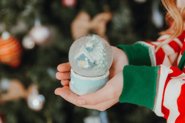 Ein kleines Mädchen im Pyjama und einer Weihnachtsmütze hält einen Weihnachtsball - eine Schneekugel. Weihnachten