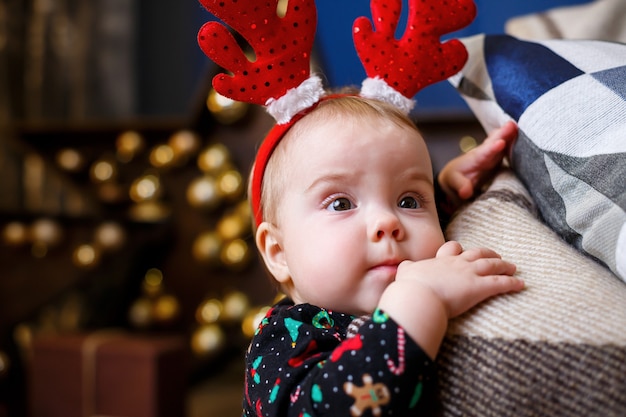 Ein kleines Mädchen im warmen Pullover sitzt unter einem Weihnachtsbaum mit Spielzeug und Geschenken mit Hörnern auf dem Kopf. Glückliche Kindheit. Silvester-Ferienatmosphäre