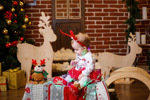 Ein kleines Mädchen im warmen Pullover sitzt unter einem Weihnachtsbaum mit Spielzeug und Geschenken mit Hörnern auf dem Kopf. Glückliche Kindheit. Silvester-Ferienatmosphäre