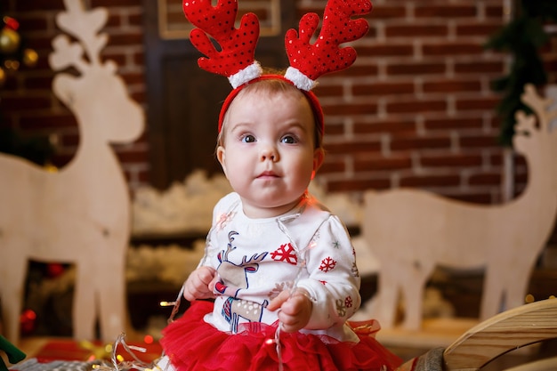 Ein kleines Mädchen im warmen Pullover sitzt unter einem Weihnachtsbaum mit Spielzeug und Geschenken mit Hörnern auf dem Kopf. Glückliche Kindheit. Silvester-Ferienatmosphäre
