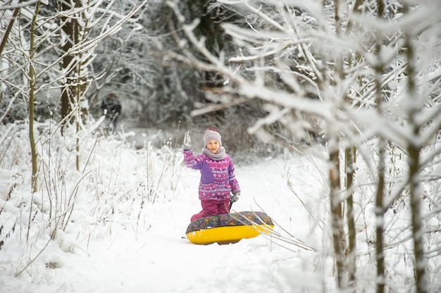 Ein kleines Mädchen im Winter in lila Kleidung und einem aufblasbaren Kreis geht auf der Straße in einem schneebedeckten Wald spazieren.