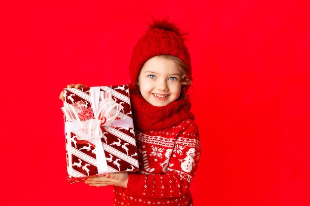 Ein kleines Mädchen in der Winterkleidung hält ein Geschenk auf einem roten Hintergrund. Neujahrskonzept, Platz für Text