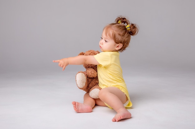 Ein kleines mädchen in einem gelben bodysuit sitzt und spielt mit einem teddybären auf weißem hintergrund