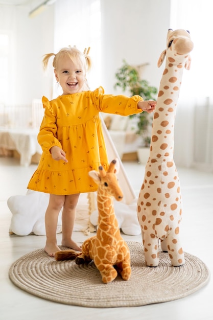 Ein kleines Mädchen in einem gelben Kleid spielt zu Hause mit einer Giraffe, die ihre Größe in einem hellen Raum misst und eine glückliche Kindheit lächelt