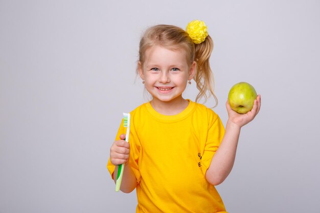 Ein kleines Mädchen in einem gelben T-Shirt hält eine Zahnbürste und einen Apfel auf weißem Hintergrund