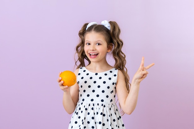 Ein kleines Mädchen in einem gepunkteten Kleid hält eine Orange und zeigt ein Häschen Vitamine für Kinder