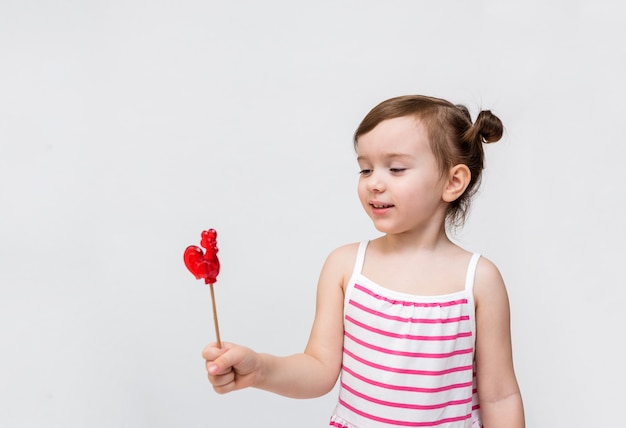 Ein kleines Mädchen in einem gestreiften Kleid mit einem Lutscher. Lutscher auf einem Stock in Form eines Hahns. Süß für Kinder