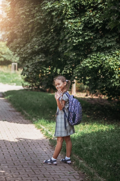 Ein kleines Mädchen in einem Kleid mit Zöpfen hält einen großen blauen Rucksack, winkt mit der Hand und geht auf der Straßengasse Erster Septembertag in der Grundschule