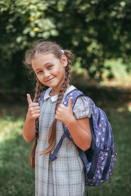 Ein kleines Mädchen in einem Kleid mit Zöpfen hält einen großen blauen Rucksack, zeigt Klasse mit der Hand und geht auf der Straßengasse spazieren Erster Septembertag in der Grundschule