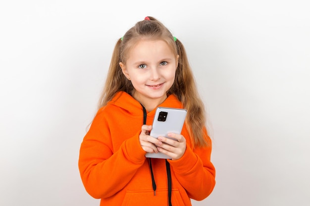 Ein kleines Mädchen in einem orangefarbenen Anzug schaut auf ihr Telefon Abhängigkeit einer Generation von Kindern von Gadgets auf weißem Hintergrund