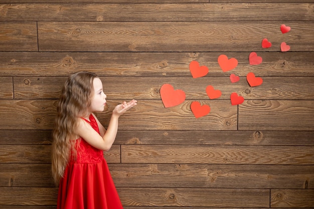 Ein kleines Mädchen in einem roten Kleid bläst Herzen von ihren Händen auf einem dunkelbraunen Holzhintergrund das Konzept des Valentinstags ein leerer Raum für Text