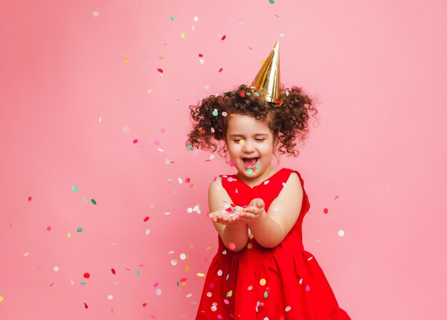 Ein kleines Mädchen in einem roten Kleid feiert ihren Geburtstag und bläst und fängt Konfetti auf einem rosa Hintergrund