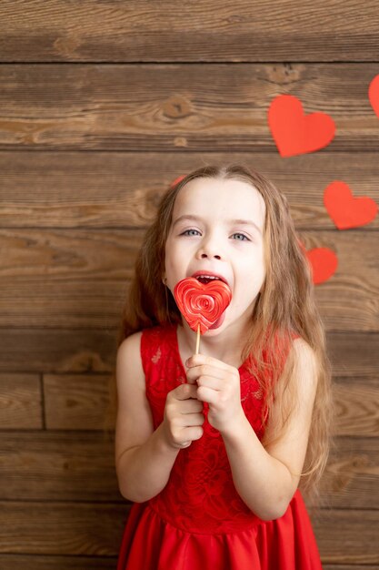 ein kleines Mädchen in einem roten Kleid leckt mit der Zunge einen großen Lutscher in Form eines roten Herzens auf einem dunkelbraunen Holzgrund mit roten Herzen das Konzept des Valentinstags ein Ort für Text