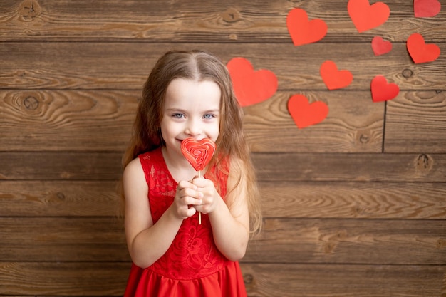 Ein kleines Mädchen in einem roten Kleid leckt mit ihrer Zunge einen großen Lutscher in Form eines roten Herzens auf einem dunkelbraunen Holzhintergrund mit roten Herzen das Konzept des Valentinstags ein Ort für Text