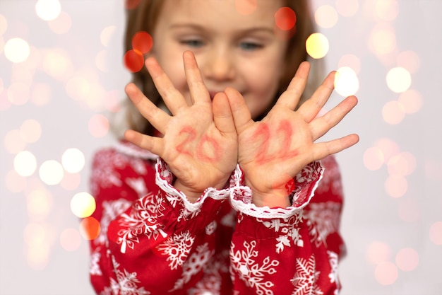 Ein kleines Mädchen in einem roten Kleid mit Schneeflocken hält ihre Handflächen vor sich. Auf den Handflächen sind rote Zahlen 2021 geschrieben
