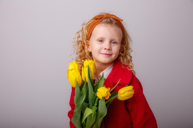 Ein kleines Mädchen in einem roten Mantel hält einen Blumenstrauß aus Frühlingstulpen auf weißem Hintergrund