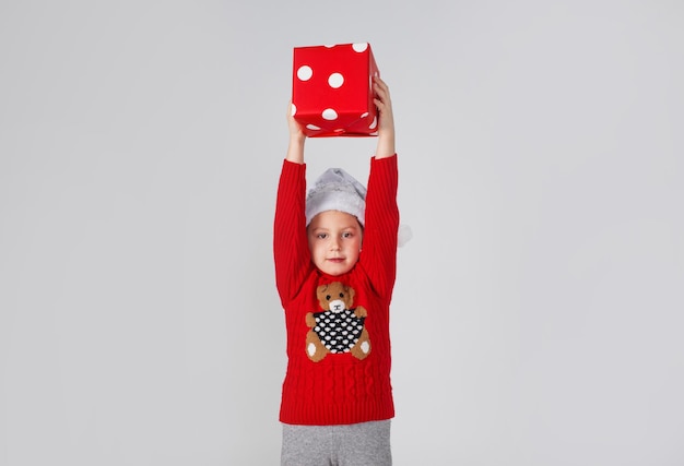 Ein kleines Mädchen in einem roten Pullover hebt eine Geschenkbox hoch, Freude ist positiv auf einem isolierten weißen Hintergrund. Weihnachten.
