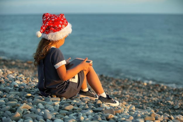 Ein kleines Mädchen in einem roten Weihnachtsmann-Hut sitzt mit einem Tablet am Meer