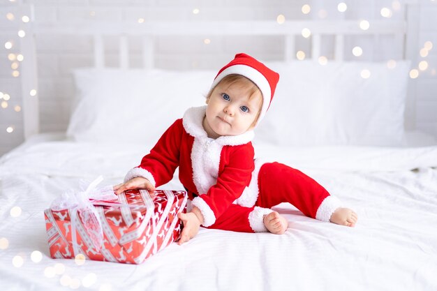 Ein kleines Mädchen in einem roten Weihnachtsmannkostüm sitzt auf einem Bett mit Weihnachtsgeschenken in Kisten