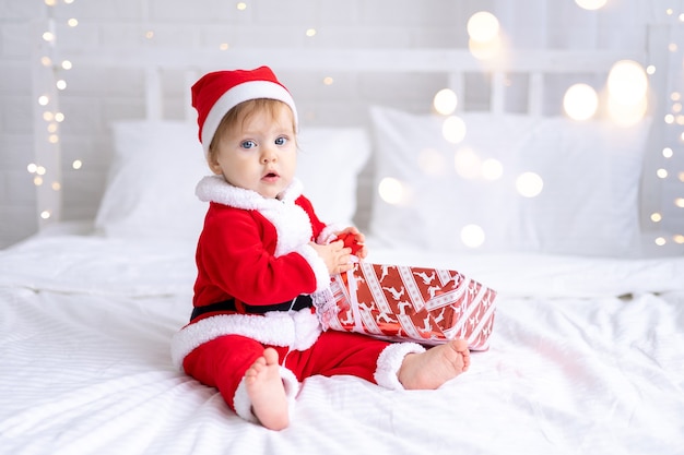 Ein kleines Mädchen in einem roten Weihnachtsmannkostüm sitzt auf einem Bett mit Weihnachtsgeschenken in Kisten