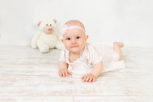 Ein kleines Mädchen in einem weißen Kleid mit einem Spielzeug liegt sechs Monate auf dem Bauch