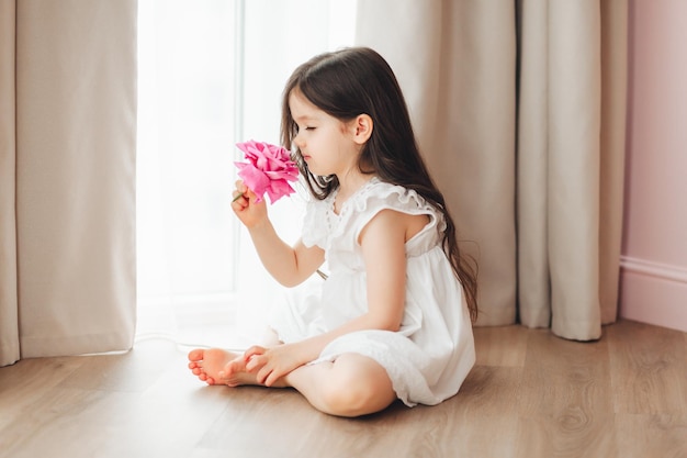 Ein kleines Mädchen in einem weißen Kleid sitzt am Fenster und hält eine Rose Ein glückliches Kind am Fenster mit einer Blume