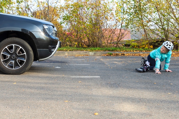 Ein kleines Mädchen in einer blauen Jacke und einem Helm auf Rollen fiel vor einem fahrenden Auto auf einer Stadtstraße