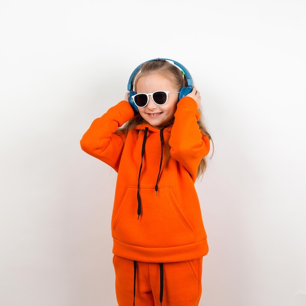 Ein kleines Mädchen in einer orangefarbenen Hoody-Sonnenbrille und blauen Kopfhörern hört Musik Porträt auf weißem Hintergrund