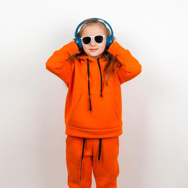 Ein kleines Mädchen in einer orangefarbenen Hoody-Sonnenbrille und blauen Kopfhörern hört Musik Porträt auf weißem Hintergrund