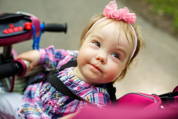 Ein kleines Mädchen in einer Schleife auf dem Kopf sitzt am Steuer eines Fahrrads. Das erste Kinderfahrrad. Sommerspaziergang mit Baby