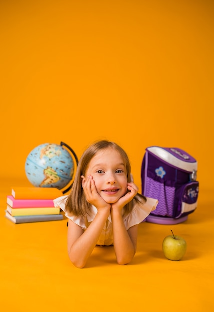 Ein kleines Mädchen in einer Schuluniform liegt auf dem Boden mit Schulmaterial auf gelbem Hintergrund mit Platz für Text