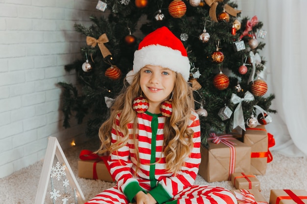 Ein kleines Mädchen in einer Weihnachtsmütze sitzt auf dem Boden neben dem Weihnachtsbaum und hält ein Geschenk in den Händen. Heiligabend.