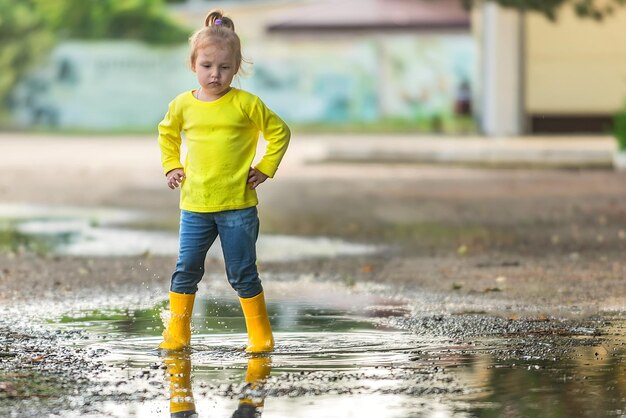 Ein kleines Mädchen in gelben Kleidern und Gummistiefeln läuft nach dem Regen auf einem Spaziergang durch die Pfützen