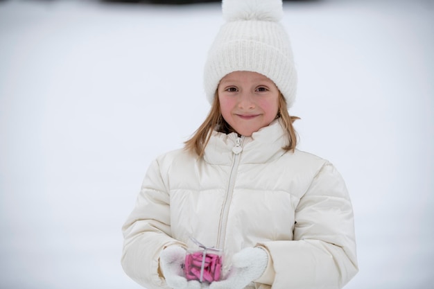 Foto ein kleines mädchen in weißer winterkleidung im schnee, das eine transparente schachtel mit süßigkeiten in form von herzen hält