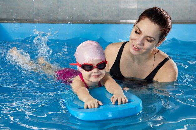 Ein kleines Mädchen lernt mit einem Poolbrett und einem Trainer im Indoor-Pool zu schwimmen