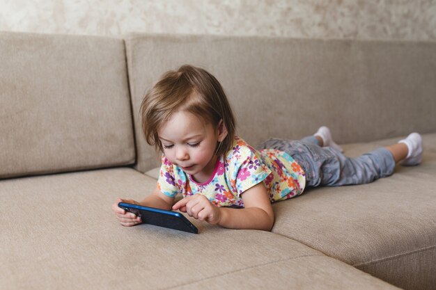 Ein kleines Mädchen liegt auf der Couch und schaut auf das Telefon