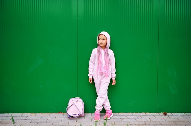 Ein kleines Mädchen mit Afro-Zöpfen posiert gegen eine grüne Wand in einem rosa Overall.