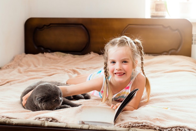 Ein kleines Mädchen mit blonden Haaren liegt zu Hause auf einem Bett, während eine Katze ein Buch liest. Bleib zuhause. Heimerziehung für Kinder.