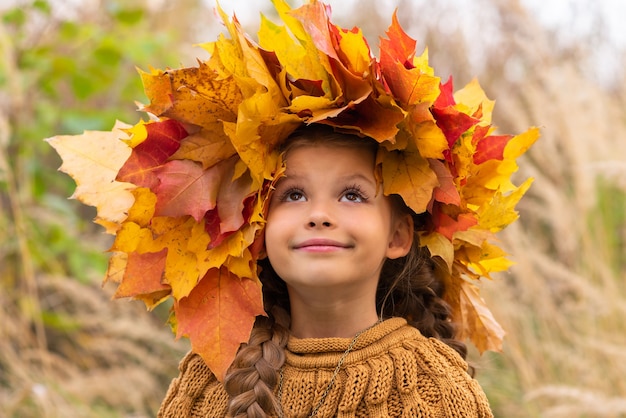 Ein kleines Mädchen mit einem Kranz aus Ahornblättern auf dem Kopf in einem warmen Pullover an einem Herbsttag.