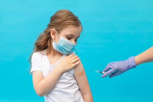 Ein kleines Mädchen mit einem Patch auf dem Arm nach der Impfung in einer medizinischen Schutzmaske auf blauem Hintergrund und die Hand einer Krankenschwester mit einer Spritze, Platz für Text, das Konzept der Impfung