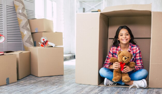 Ein kleines Mädchen mit einem Teddybär versteckt sich in einem Pappkarton und spielt wahrscheinlich mit ihren Eltern in ihrer geräumigen Wohnung Verstecken