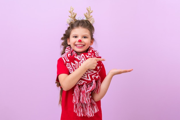 Ein kleines Mädchen mit Hirschohren auf dem Kopf und aufgemalter Nase weist auf Ihre Werbung hin. Ein Mädchen wartet auf Weihnachten auf einem lila isolierten Hintergrund.