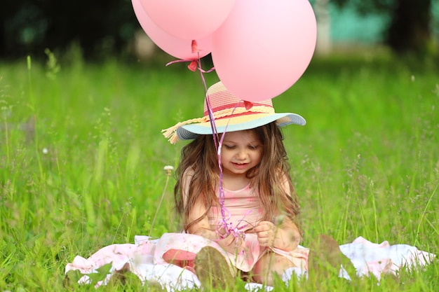Ein kleines Mädchen mit Hut, das im Sommer mit Luftballons auf dem Gras sitzt. Foto in hoher Qualität