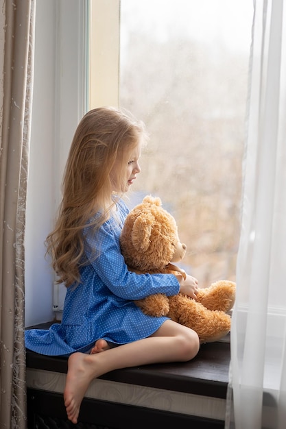 Ein kleines Mädchen mit langen blonden Haaren sitzt mit einem Spielzeug auf der Fensterbank und schaut aus dem Fenster auf die Straße. Dies ist eine traurige Stimmungskrankheit oder Isolation